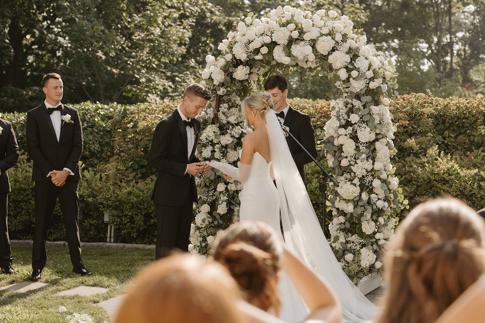 Groom places a ring on the bride's finger under a white rose arch during the ceremony.